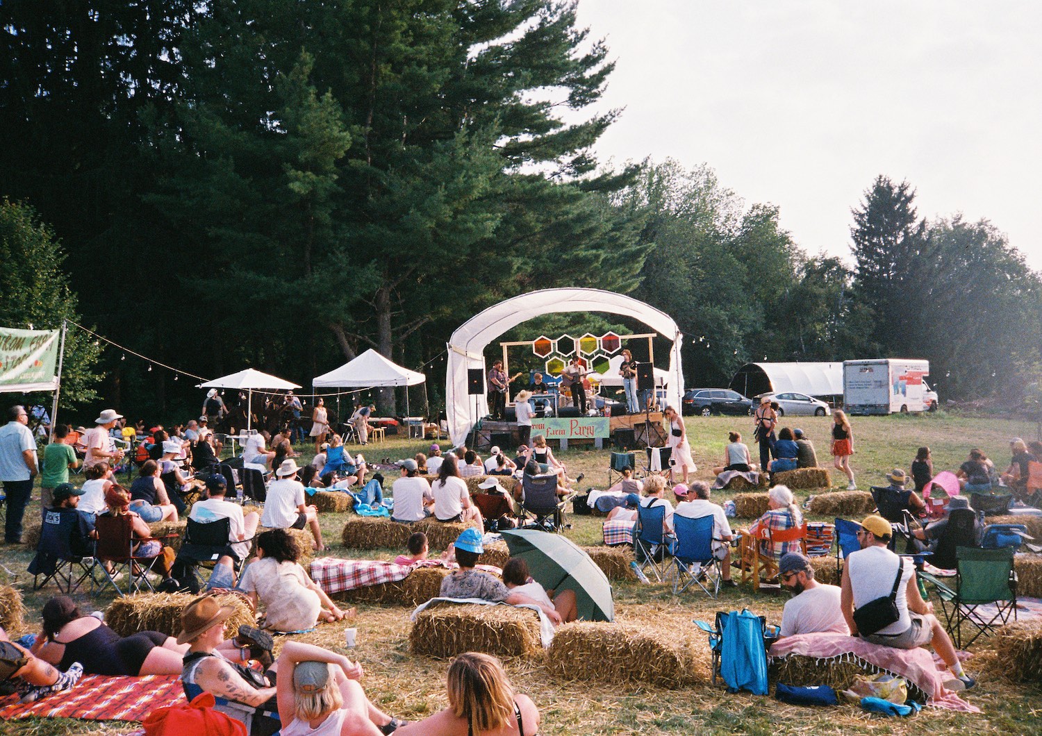Attendees sitting on haybales in a grassy field watch an artist perform at Avrom Farm Party.