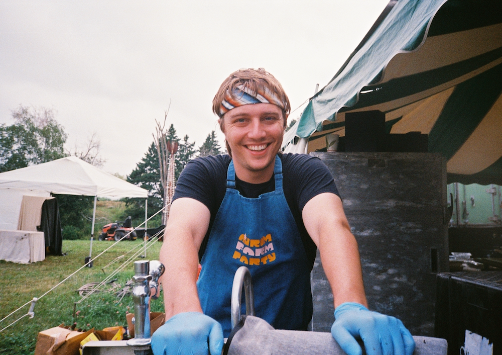 A festival employee smiles wearing a denim apron and bandana tied around his forehead while leaning on a sink.