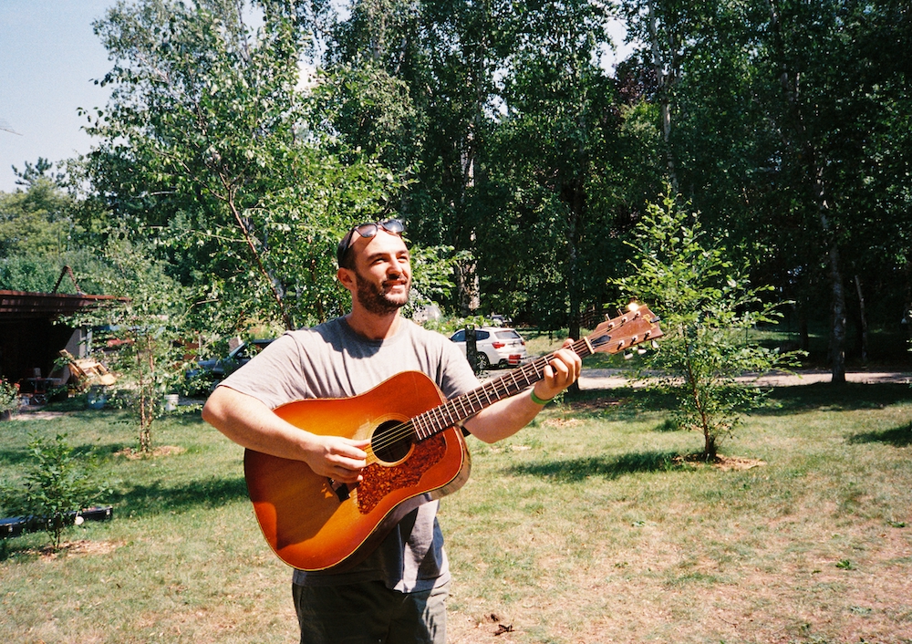 A festival artist plays acoustic guitar among trees.